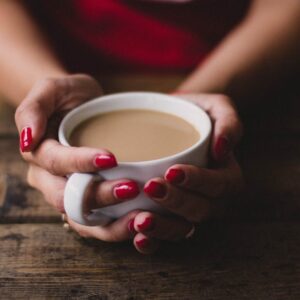 Warm indoor scene of a woman holding a steaming cup of coffee with red nails.